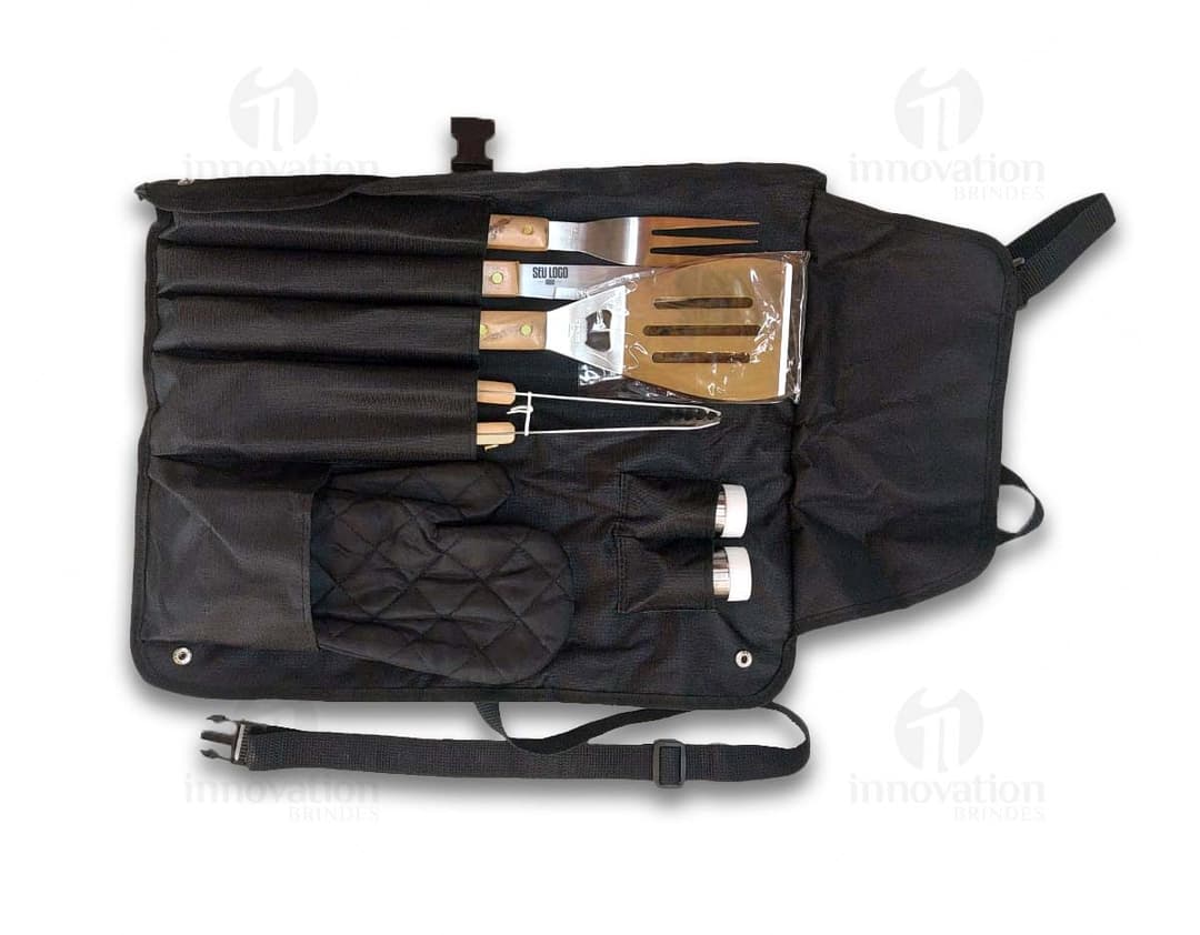 Homem sorrindo, em pé, segurando um kit de churrasco completo com avental preto e utensílios de metal, como faca, garfo e espátula. Ele veste uma jaqueta de couro e possui uma mochila nas costas.