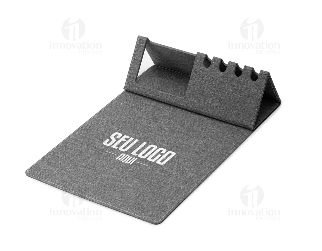 Mouse pad de couro marrom aberto em cima de uma mesa de escritório, ao redor estão um notebook preto, uma bolsa de couro, chaves, um cartão de crédito, uma carteira, um bloco de notas, um teclado preto e um mouse preto.