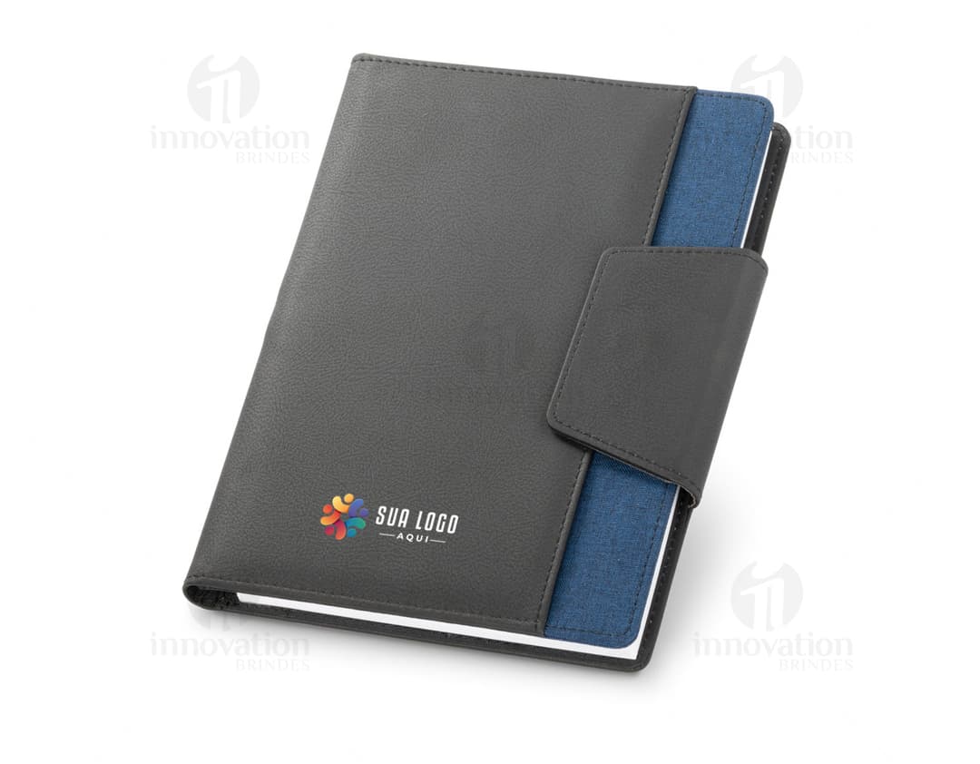 Notebook A5 com capa de couro sintético preto aberto sobre uma mesa de escritório. O caderno está vazio, com um teclado de computador ao lado, um estojo de negócios preto, um suporte para papel, discos magnéticos e notas espalhadas.