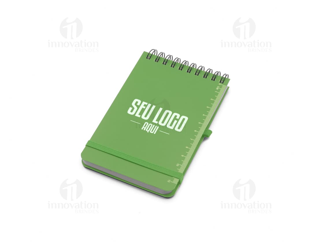 Mini caderno em 3D, com capa preta e espiral, aberto e vazio. O caderno está sobre uma mesa de escritório, ao lado de um notebook, um livro e um papel em branco. A imagem transmite a ideia de organização, estudo e trabalho.