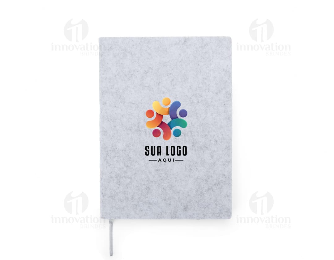 caderno A5 de feltro preto, sem pauta, aberto em uma página em branco. Ao lado, um notebook e um envelope preto. No fundo, livros e objetos de escritório.