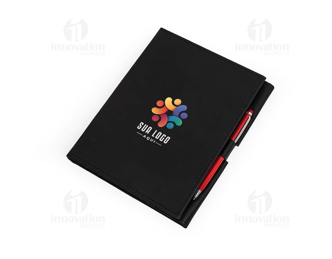 Caderneta com suporte na cor rosa, feita de material reciclado e com elástico para fechamento. O suporte é acoplado na capa da caderneta, permitindo que o usuário possa encaixar uma caneta ou lápis.