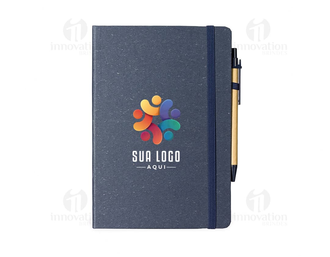 Caderneta de fibra de arroz com caneta em fundo branco. O caderno é marrom claro e possui detalhes em preto na capa. A caneta é de cor azul e está ao lado da caderneta.