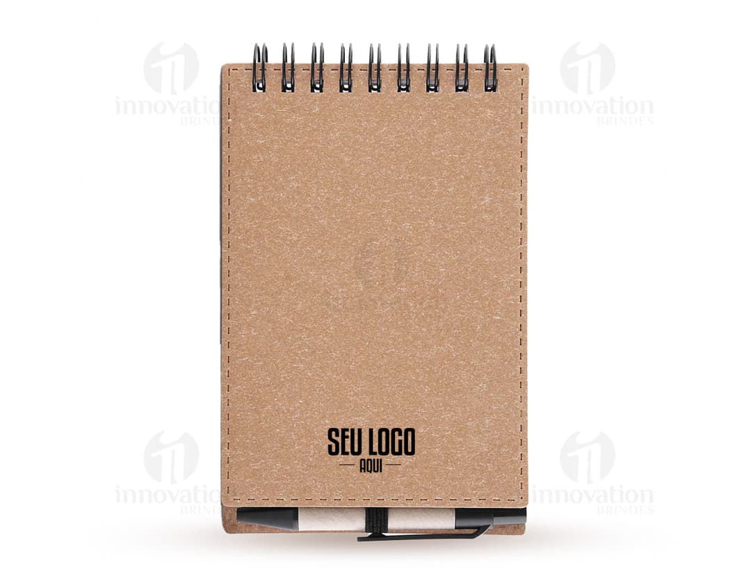 Mini caderno de anotações em papel em branco, com capa marrom de cartão. Acompanha uma caneta e está em cima de um notebook aberto em um escritório, ao lado de uma prancheta e caixa de papelão. A imagem possui um estilo vintage e grunge, com um ar antigo e de memória pessoal.